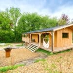 La photo montre une charmante cabane en bois sur pilotis, nichée en pleine nature, avec une large terrasse en bois qui offre un espace extérieur accueillant. Devant la cabane, deux chaises longues colorées sont installées sur un sol recouvert de copeaux de bois, invitant à la détente. À proximité, un foyer moderne en métal, de type barbecue plancha, est installé, parfait pour des moments conviviaux de cuisine en plein air. L’ensemble est entouré d’un cadre verdoyant, avec des arbres et des prairies, offrant un véritable havre de paix en pleine nature.