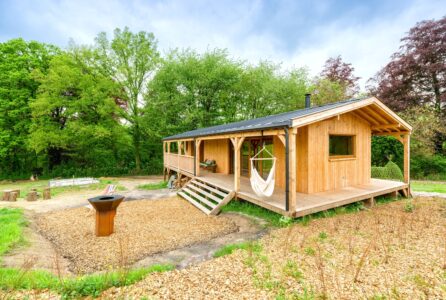 La photo montre une charmante cabane en bois sur pilotis, nichée en pleine nature, avec une large terrasse en bois qui offre un espace extérieur accueillant. Devant la cabane, deux chaises longues colorées sont installées sur un sol recouvert de copeaux de bois, invitant à la détente. À proximité, un foyer moderne en métal, de type barbecue plancha, est installé, parfait pour des moments conviviaux de cuisine en plein air. L’ensemble est entouré d’un cadre verdoyant, avec des arbres et des prairies, offrant un véritable havre de paix en pleine nature.