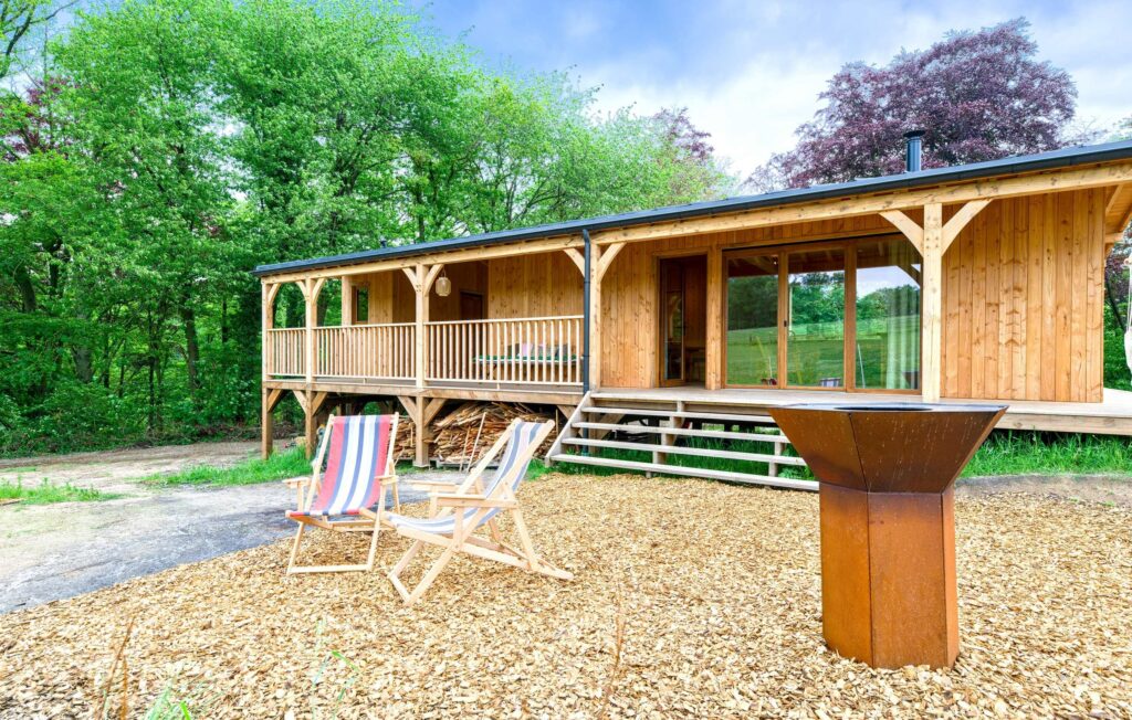 Cabane en bois sur pilotis avec terrasse, entourée de verdure. Devant, deux chaises longues colorées et un barbecue plancha en métal moderne, parfait pour des moments conviviaux en plein air, dans un cadre naturel et apaisant