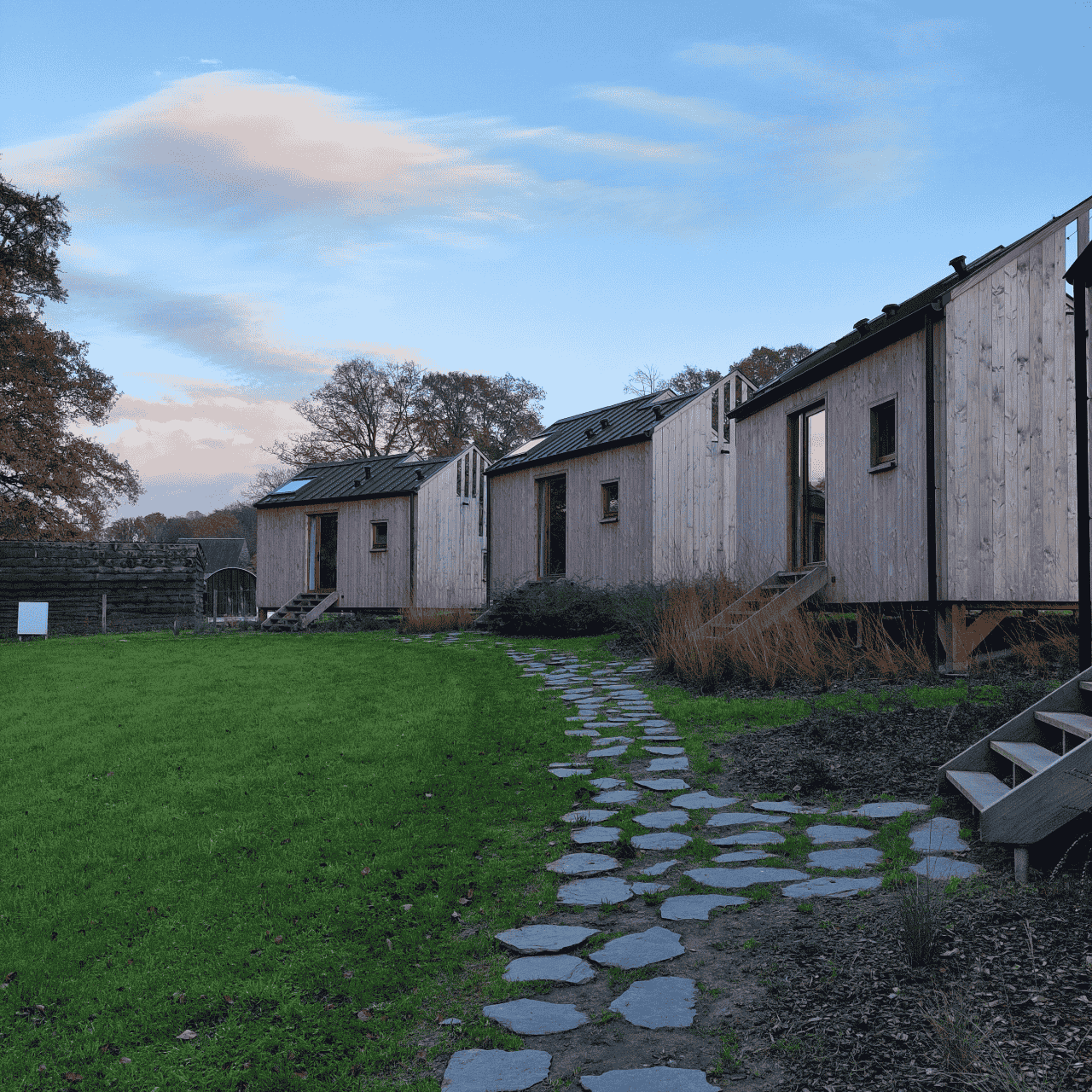 Un alignement de cabanes en bois sur pilotis, entourées de verdure et de plantes sauvages, avec un chemin de pierres serpentant à travers une pelouse bien entretenue. À l'arrière-plan, des arbres aux teintes automnales s'étendent sous un ciel bleu parsemé de nuages légers, créant une atmosphère paisible et naturelle.